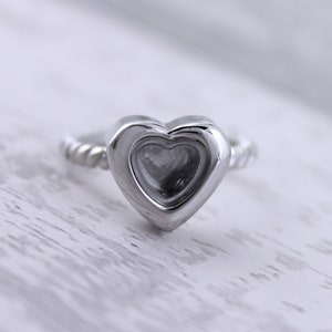 Puede incluir: Un anillo de plata con un diseño en forma de corazón. El corazón es hueco y tiene un corazón más pequeño en su interior.