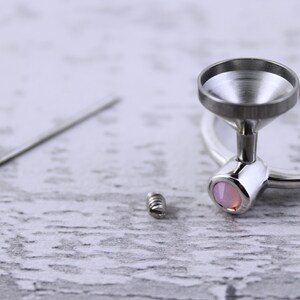 May include: A silver ring with a funnel-shaped top and a pink stone set in the base. The ring is shown with a small silver screw and a silver pin.