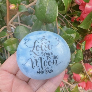 May include: A blue painted rock with the words "Love you to the moon and back" written in black calligraphy. The rock is decorated with a crescent moon and stars.