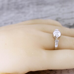 May include: A silver ring with a round, raised center stone and a delicate band. The ring is set on a hand, showcasing its design and details. The ring's design is simple and elegant.