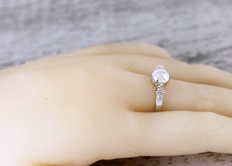 May include: A silver ring with a round, raised center stone and a delicate band. The ring is set on a hand, showcasing its design and details. The ring's design is simple and elegant.