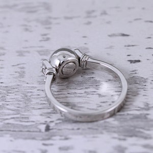May include: A silver ring with a round, domed top and a simple band. The ring has a unique design with decorative elements on either side of the domed top. The ring is set against a white, textured background.
