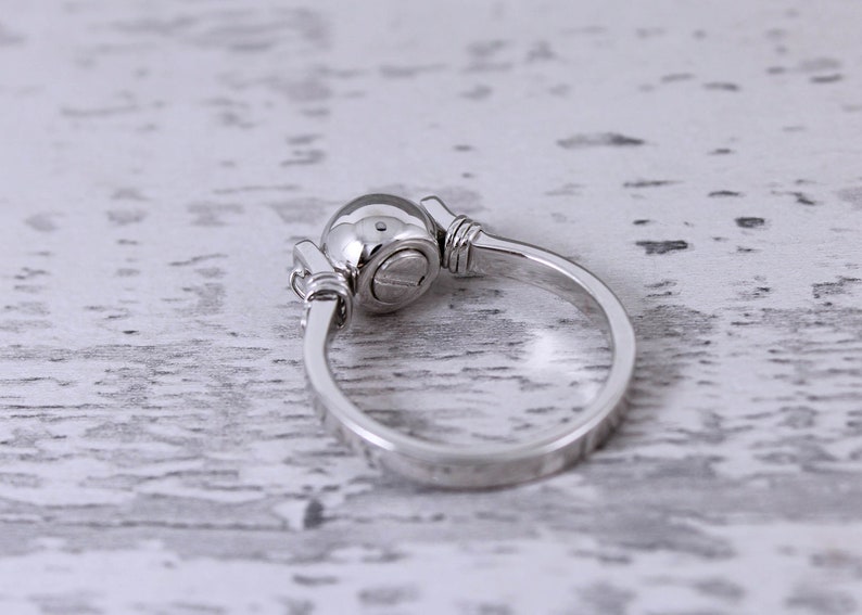 May include: A silver ring with a round, domed top and a simple band. The ring has a unique design with decorative elements on either side of the domed top. The ring is set against a white, textured background.