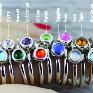 May include: A set of 14 silver rings with different colored gemstones. The gemstones are garnet, amethyst, aquamarine, crystal, emerald, light amethyst, light siam, peridot, sapphire, rose, opal, topaz, blue zircon, and pink opal.
