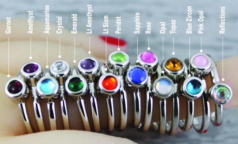 May include: A set of 14 silver rings with different colored gemstones. The gemstones are garnet, amethyst, aquamarine, crystal, emerald, light amethyst, light siam, peridot, sapphire, rose, opal, topaz, blue zircon, and pink opal.