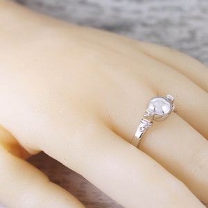 May include: A silver ring with a round, pearl-like center stone. The ring's band features delicate, decorative detailing. The ring is shown on a person's finger, highlighting its design and craftsmanship. A piece of jewelry.