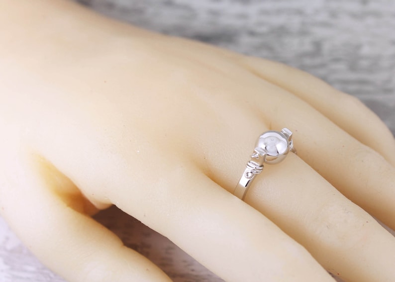 May include: A silver ring with a round, pearl-like center stone. The ring's band features delicate, decorative detailing. The ring is shown on a person's finger, highlighting its design and craftsmanship. A piece of jewelry.