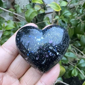 May include: A heart-shaped, black stone with a speckled, galaxy-like design in shades of blue, purple, and white. The polished stone is held in a hand, with a natural green background of leaves and branches.