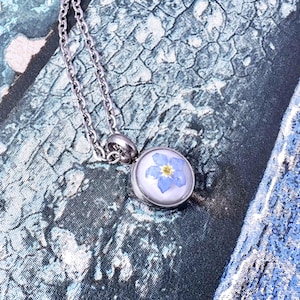 Op de afbeelding: Een zilverkleurige ketting met een ronde hanger met een blauwe vergeet-mij-nietje bloem ingekapseld in heldere hars. De ketting is delicaat en de hanger is geplaatst tegen een blauw en wit getextureerde achtergrond.