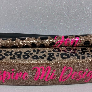 May include: A glittery pen and pen holder set. The pen and holder are covered in a gold glitter leopard print design with black spots. The pen has the word "Hope" in pink. The pen holder has the words "Inspire Mi Design" in pink.