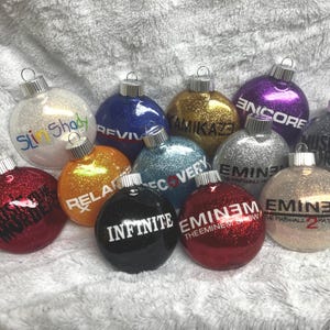 May include: A selection of glittery Christmas baubles in diverse colours, including red, gold, and silver. Each bauble displays text like "Slim Shady", "Kamikaze", and "Encore". The baubles have silver metal caps.