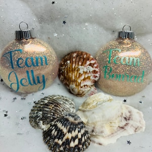 May include: Two glittery, pearlescent Christmas ornaments with the words "Team Jelly" and "Team Bread" written in teal on them. The ornaments are surrounded by seashells.