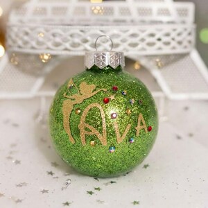 May include: A round green glitter Christmas ornament featuring a gold silhouette of a fairy and the name AVA in gold lettering. The ornament is decorated with small colorful rhinestones and has a silver metal cap with a hanging loop at the top.