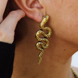 May include: Gold snake earrings with a textured surface. The earrings are long and dangle from the earlobe.