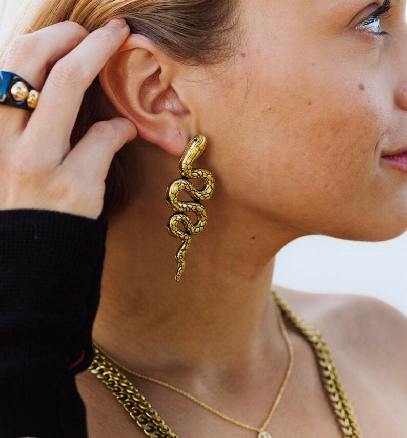 K&ouml;nnte beinhalten: Goldene Schlangenohrringe mit strukturierter Oberfl&auml;che. Die Ohrringe sind lang und h&auml;ngen vom Ohrl&auml;ppchen.