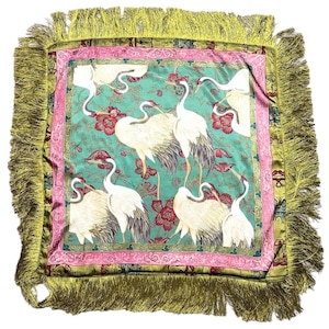 May include: A square decorative pillow featuring a central teal panel with white crane birds and red floral patterns. The design is framed by a pink border and an outer olive green border, finished with long, matching olive green fringe on all sides.