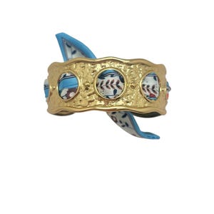 May include: Gold-toned cuff bracelet with a textured surface and three circular insets. Each inset features a patterned fabric in shades of blue, white, and brown. A blue and white patterned scarf is threaded through the bracelet.
