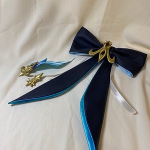 Puede incluir: Una pajarita azul oscuro con forro azul claro y un acento decorativo dorado. La pajarita tiene dos colas largas que también están forradas en azul claro.