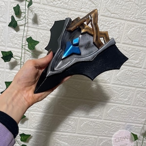 May include: A stylized mask featuring a black base, a silver metallic center, and a bright blue diamond-shaped accent. The upper portion includes a textured bronze-colored structure with angular, branch-like protrusions extending upward.