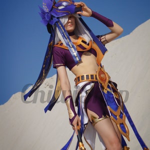May include: A woman in a purple and gold costume with a blue and gold headpiece, holding a staff with a gold and brown handle. She is standing in a desert setting.
