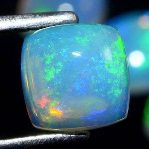May include: A light blue opal gemstone with green and blue flashes of color. The gemstone is cut into a square shape with rounded edges.