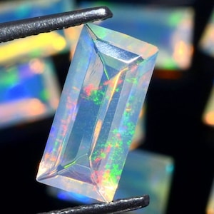 Puede incluir: Una piedra preciosa de ópalo rectangular, facetada y transparente con colores arcoíris iridiscentes. La piedra preciosa está sujeta por unas pinzas.