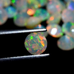 May include: A round, white opal gemstone with green and orange flashes of color. The opal is held in tweezers against a black background.