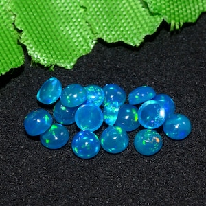 May include: A group of 14 blue opal cabochons, each with a slightly different shade of blue and a hint of green. The opals are arranged in a loose pile on a black surface.