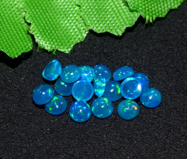 May include: A group of 14 blue opal cabochons, each with a slightly different shade of blue and a hint of green. The opals are arranged in a loose pile on a black surface.