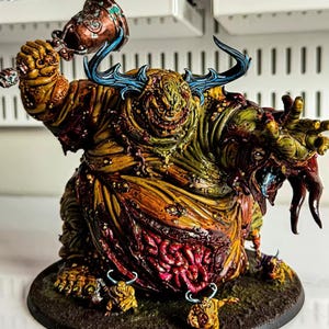May include: A hand-painted miniature figurine of a large, corpulent creature with mottled yellow, green, and brown skin. It features prominent blue horns, a wide grin, and a bell held aloft. The torso displays exposed red viscera, and two smaller figures stand at its base on a circular textured platform.