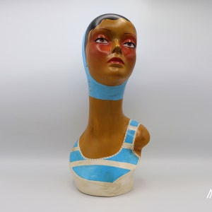 Może przedstawiać: Ceramiczny popiersie kobiety w niebiesko-białym pasiastym kostiumie kąpielowym i niebieskim czepkiem kąpielowym. Popiersie jest namalowane w realistycznym stylu i ma vintage'owy wygląd.