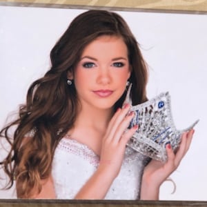May include: A young woman with long brown hair is holding a silver crown with blue jewels. She is wearing a white dress with a beaded neckline.