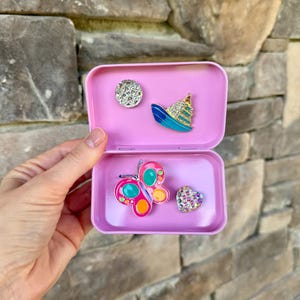 May include: A light pink metal storage tin containing four decorative pins: a circular rhinestone pin, a cruise ship pin with blue and gold accents, a colorful butterfly pin with pink and teal enamel, and a small heart-shaped rhinestone pin.