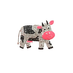 May include: A black and white cow brooch with pink accents and rhinestones. The cow is standing on all four legs and has a smiling face.