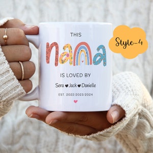 May include: White ceramic mug with a rainbow design and the text "This Nana is loved by Sera Jack Danielle Est. 2022/2023/2024". The mug is being held by a person's hand.