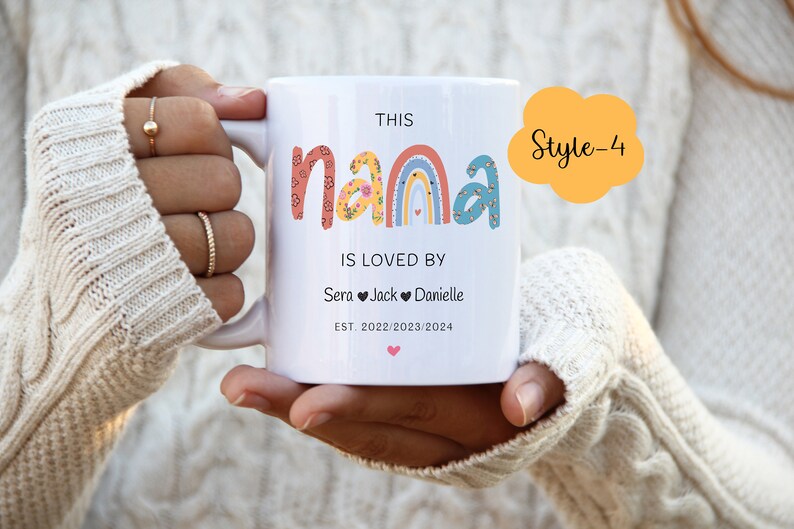 May include: White ceramic mug with a rainbow design and the text "This Nana is loved by Sera Jack Danielle Est. 2022/2023/2024". The mug is being held by a person's hand.