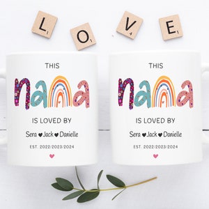 May include: Two white ceramic mugs with a rainbow design and the text "This Nana is loved by" followed by names and dates. The mugs are on a white surface with wooden letter tiles spelling out "LOVE".
