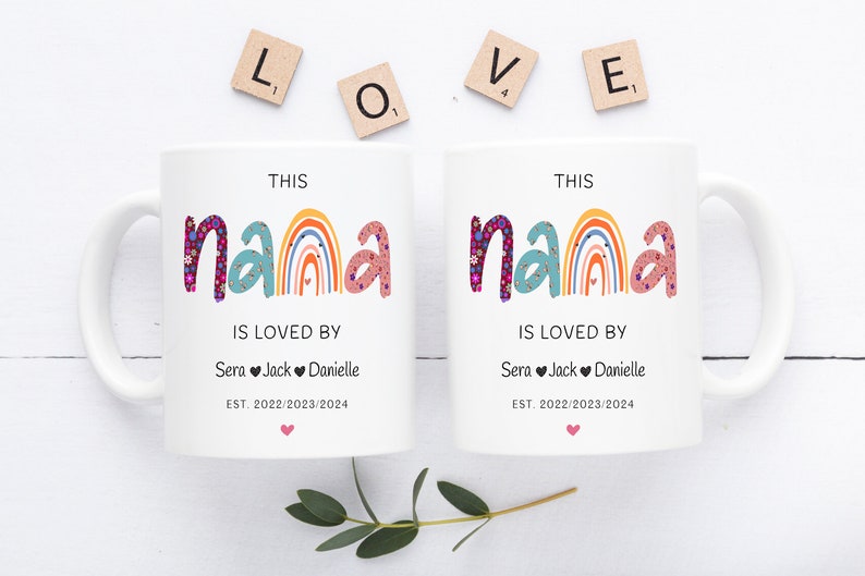 May include: Two white ceramic mugs with a rainbow design and the text "This Nana is loved by" followed by names and dates. The mugs are on a white surface with wooden letter tiles spelling out "LOVE".