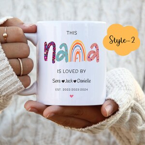 May include: White ceramic mug with a rainbow design and the text "This Nana is loved by Sera Jack Danielle Est. 2022/2023/2024". The mug has a pink heart at the bottom and a yellow cloud with the text "Style-2" in the top right corner.