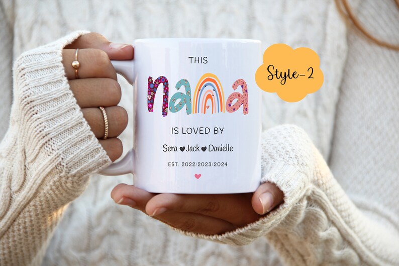 May include: White ceramic mug with a rainbow design and the text "This Nana is loved by Sera Jack Danielle Est. 2022/2023/2024". The mug has a pink heart at the bottom and a yellow cloud with the text "Style-2" in the top right corner.