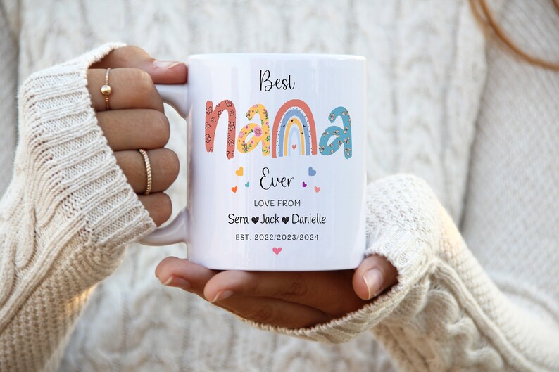 May include: White ceramic mug with a colorful rainbow design and the text "Best Nana Ever" with hearts. The text "Love from Sera Jack Danielle" and "EST. 2022/2023/2024" is also included.