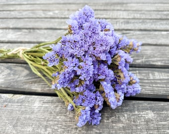 May include: A bundle of dried purple statice flowers tied with twine. The flowers have a delicate, textured appearance and are arranged against a weathered, gray wooden surface. The stems are green and the overall aesthetic is natural and rustic.