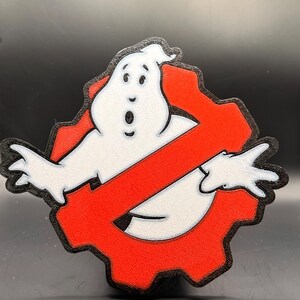 May include: A red and black Ghostbusters logo with a white ghost in the center. The ghost has a surprised expression and is wearing a black outline.