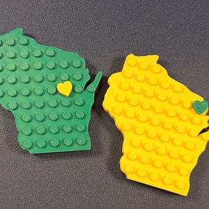 May include: Two Wisconsin-shaped coasters, one green and one yellow, each with a heart accent. The coasters have a textured surface with circular bumps, resembling building blocks. The yellow heart is on the green coaster, and the green heart is on the yellow coaster.