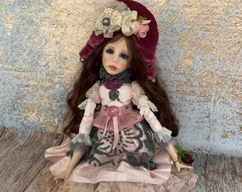 Könnte beinhalten: Handgefertigte Puppe mit langen braunen Haaren, gekleidet in einem rosa-grauen Kleid mit Spitzendetails. Die Puppe trägt einen bordeauxfarbenen Samthut mit Blumen und sitzt auf einer Holzoberfläche.