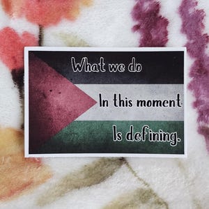 May include: A sticker featuring a black, red, and green flag design. The text on the sticker reads "What we do in this moment is defining."