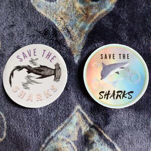 May include: Two round stickers with the text "Save the Sharks" and images of sharks. The sticker on the left features a hammerhead shark in black and white. The sticker on the right features a grey shark with a rainbow background.