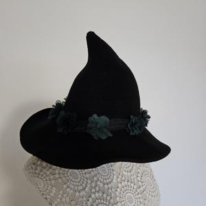Könnte beinhalten: Ein schwarzer Hexenhut mit breiter Krempe und spitzer Krone. Der Hut ist mit einem dunkelgrünen Blumenband verziert. Der Hut wird auf einer weißen Spitzenform präsentiert.