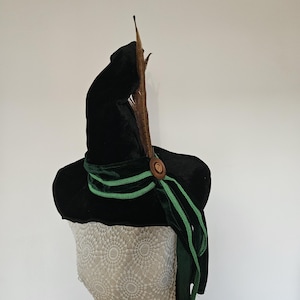 Pode incluir: Um chapéu de veludo preto com aba larga, adornado com uma fita de veludo verde e um botão de madeira decorativo. Uma pena marrom está presa no topo. O chapéu é exibido em um manequim.