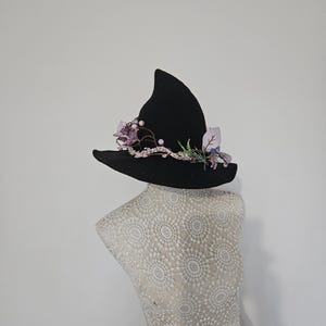 Peut inclure: Un chapeau de sorcière noir avec une couronne pointue, orné de fleurs violettes, de perles et de verdure. Le chapeau est présenté sur un mannequin. Le bord est large et légèrement incurvé.